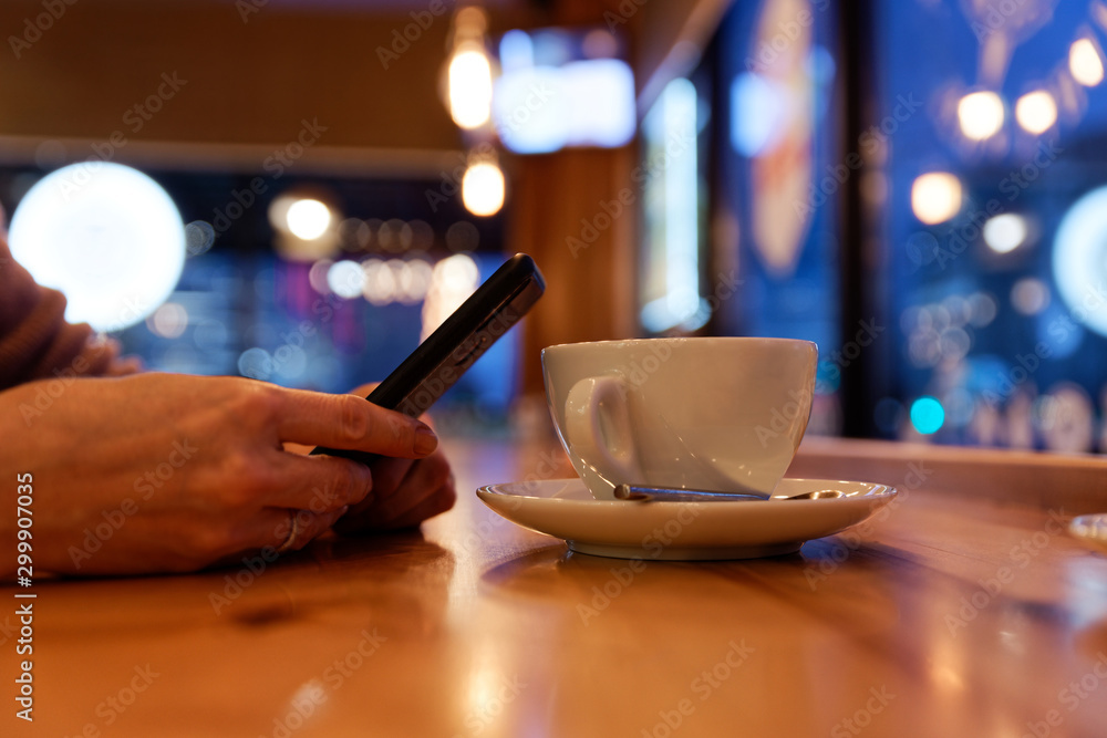 Cup of coffee or tea on the wooden table and human hands holding a smartphone against blurred background. Evening or night in a cafe.