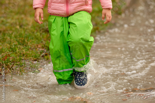 Front view of low section of a child girl in green waterproof pants and rubber boots walking through a huge rain puddle in the forest on a rainy autumn day in October in Germany
