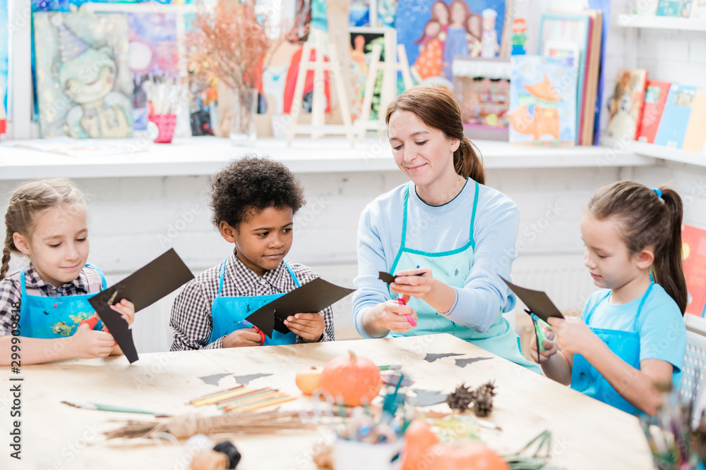 Intercultural schoolkids and their teacher in aprons cutting out halloween bats Intercultural schoolkids and their teacher in aprons cutting out halloween bats