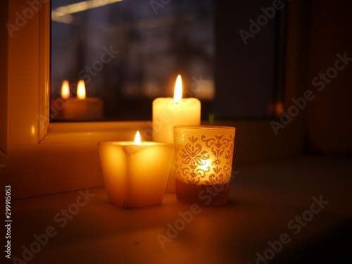 Closeup photo of three lit candles - two paraffin wax candles and one tealight in a glass candle holder with glittery golden ornament, standing on a windowsill late in the evening.