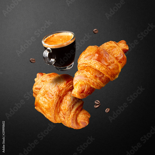 Breakfast or coffee house concept. Espresso in glass cup, croissants and beans coffee flying or falling in air on black background.