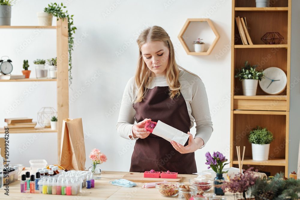 Blonde female in apron putting handmade soap bar into paper packet