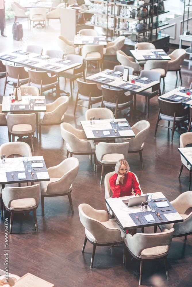 Portrait of gorgeous lady with blonde hair wearing red jumpsuit, looking at the screen of her laptop while sitting in an empty restaurant. Successful beautiful woman concept. Vertical shot. Top view Portrait of gorgeous lady with blonde hair wearing red jumpsuit, looking at the screen of her laptop while sitting in an empty restaurant. Successful beautiful woman concept. Vertical shot. Top view