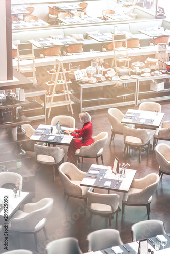 Portrait of gorgeous lady with blonde hair wearing red jumpsuit, looking at the screen of her laptop while sitting in an empty restaurant. Successful beautiful woman concept. Vertical shot. Top view