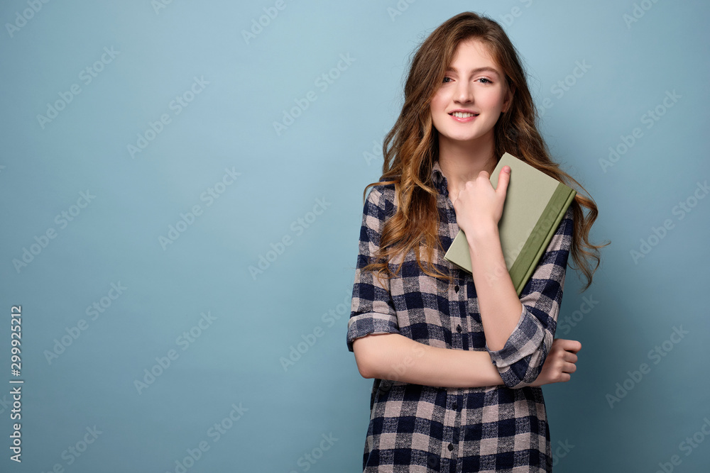A young girl in a plaid shirt stands on a blue background with a book and looks joyfully at the camera. - obrazy, fototapety, plakaty 
