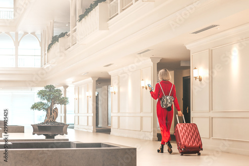 Full-length portrait of gorgeous lady with blonde hair wearing red jumpsuit entering in the hotel lobby with a backpack and a suitcase. Business trip concept. Horizontal shot. Back view