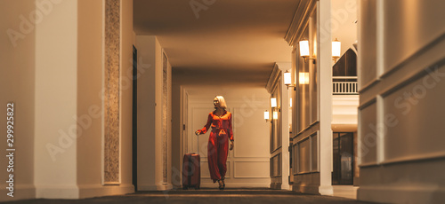 Full-length portrait of gorgeous lady with blonde hair wearing red jumpsuit walking along the hall in hotel with a suitcase. Business trip concept. Horizontal shot. Front view