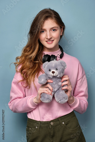 A girl in a pink sweater with headphones on her neck stands on a blue background and holds a bear toy and smiles.