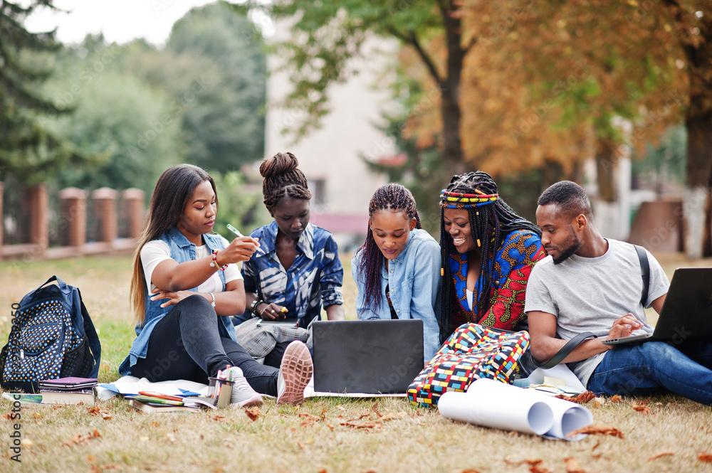 Group of five african college students spending time together on campus at university yard. Black afro friends sitting on grass and studying with laptops. - obrazy, fototapety, plakaty 