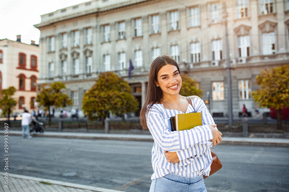 Portrait of female college student smiling at camera. Beautiful young woman with backpack and books outdoors. College student carrying lots of books in college campus.