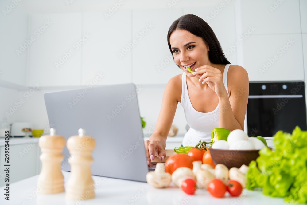 Portrait of positive cheerful housewife use her laptop searching cookery information watch culinary movies want prepare organic meal wear white singlet in kitchen house indoors Portrait of positive cheerful housewife use her laptop searching cookery information watch culinary movies want prepare organic meal wear white singlet in kitchen house indoors