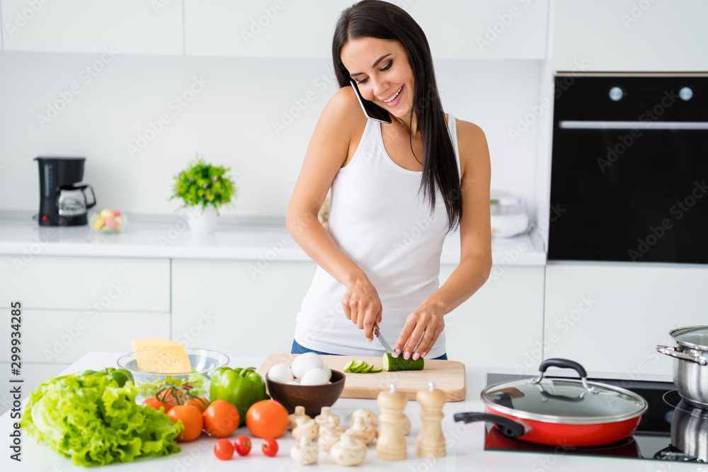 Portrait of positive cheerful girl relax weekends cooking domestic dinner supper meal speaking on cellphone with her friend cut ingredient green cucumber on chopping board in kitchen white house