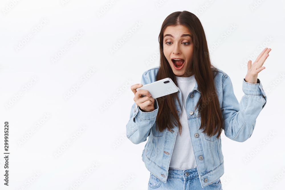 Impressed, excited and thrilled young girl shouting at smartphone astonished, raising hand up triumphing, holding mobile phone horizontally, stare screen amazed, white background Impressed, excited and thrilled young girl shouting at smartphone astonished, raising hand up triumphing, holding mobile phone horizontally, stare screen amazed, white background