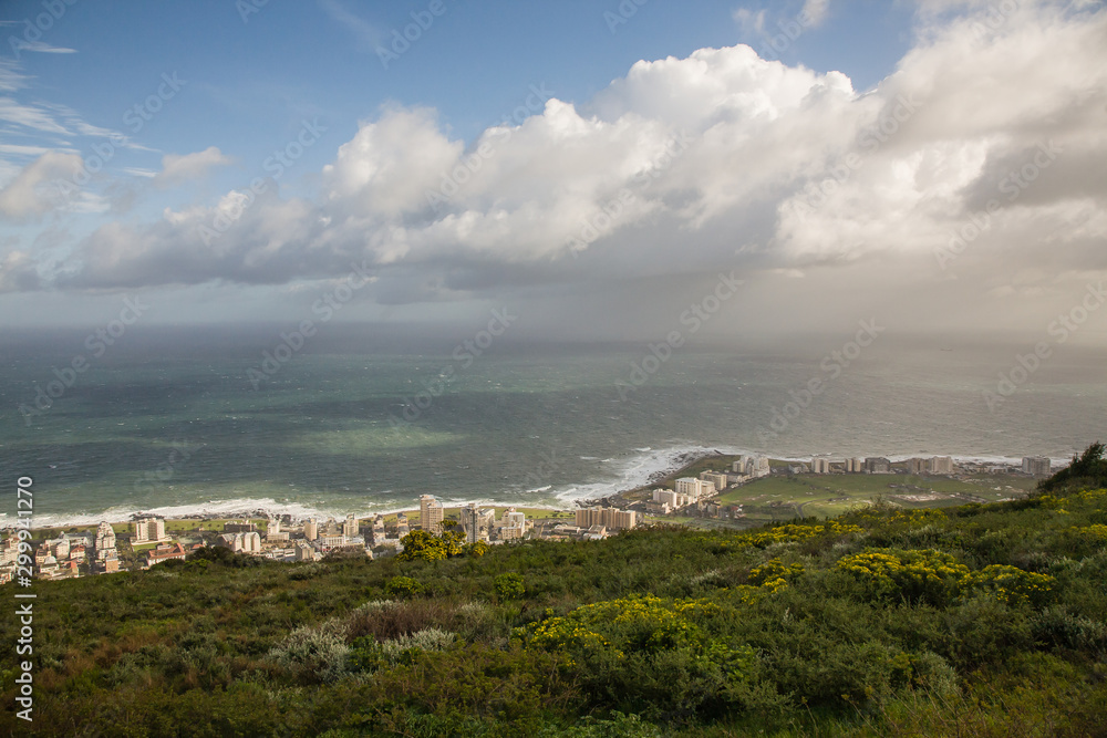 At the foot of Table Mountain, the sky over the coast of Cape Town. A cloud caught on top of a mountain. Rays of the sun illuminate the shore and buildings.