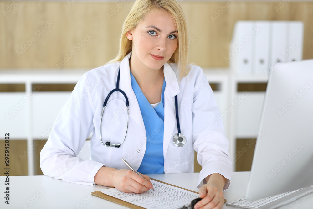 Doctor woman at work while sitting at the desk in hospital or clinic. Blonde cheerful physician filling up medical records form. Data and best service in medicine and healthcare Doctor woman at work while sitting at the desk in hospital or clinic. Blonde cheerful physician filling up medical records form. Data and best service in medicine and healthcare