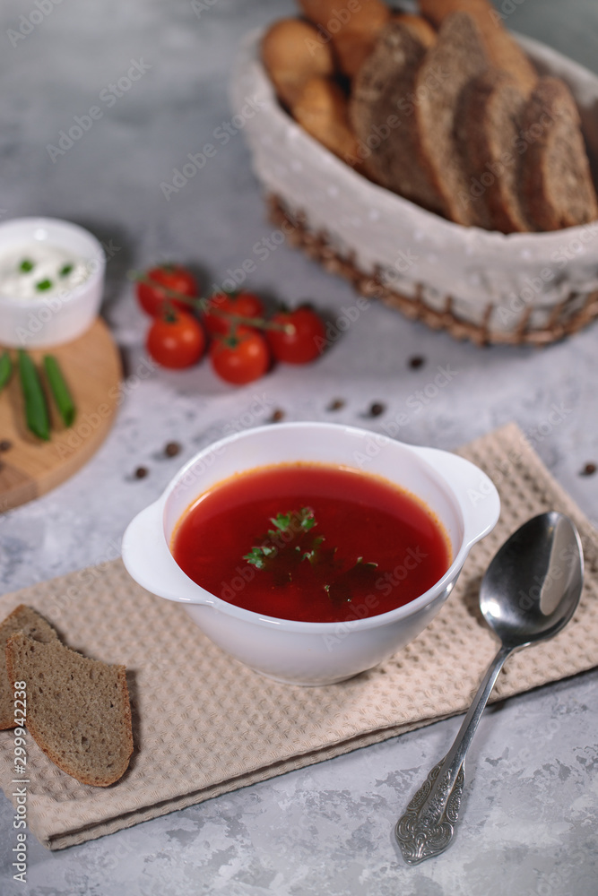 Tasty and hearty dinner. A plate with borsch on the table, next to the board are vegetables, parsley, dill, green onions, garlic, chili peppers, cherry tomatoes, and bread. Tasty and hearty dinner. A plate with borsch on the table, next to the board are vegetables, parsley, dill, green onions, garlic, chili peppers, cherry tomatoes, and bread.