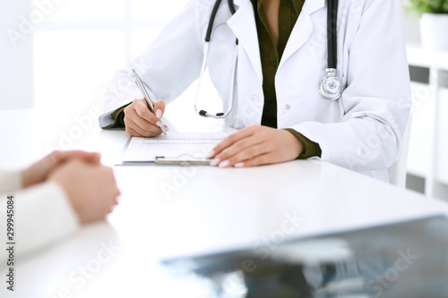 Woman doctor and patient sitting and talking at medical examination at hospital office, close-up. Therapist filling up medication history records. Medicine and healthcare concept