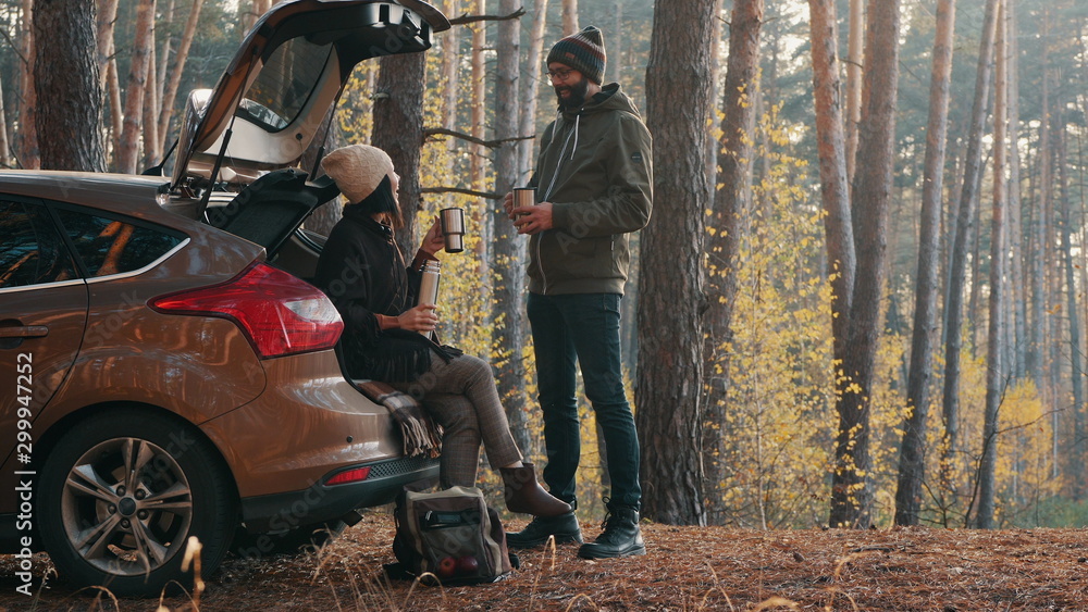 Happy smiling couple of travelers drink coffee or tea with a thermos standing near the car in the autumn forest - obrazy, fototapety, plakaty 