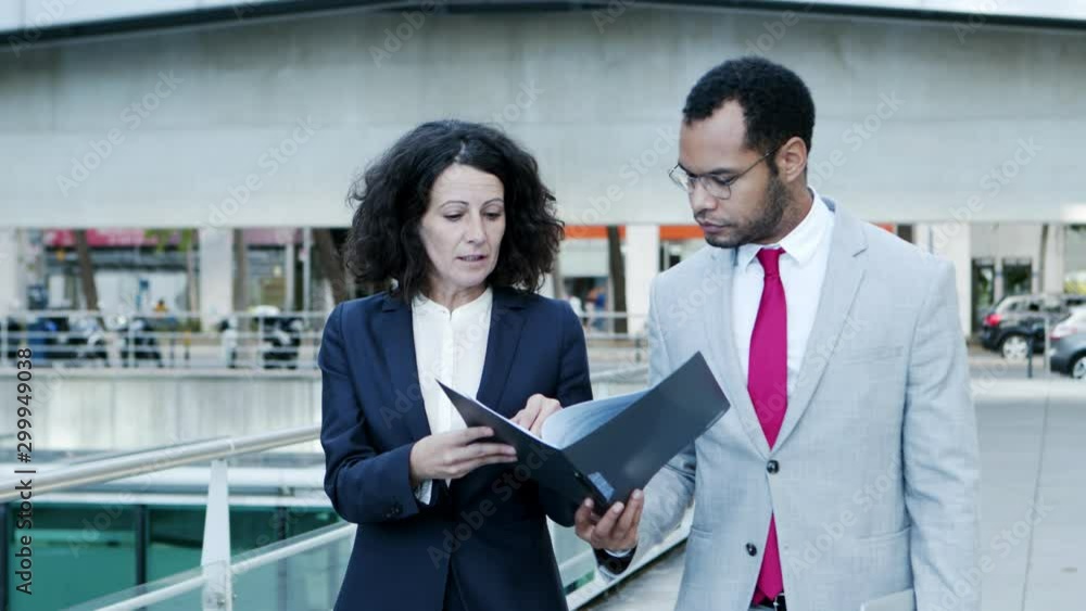 Coworkers discussing documents on street. Professional multiethnic businessman and businesswoman in formal wear walking and talking about papers. Cooperation concept Coworkers discussing documents on street. Professional multiethnic businessman and businesswoman in formal wear walking and talking about papers. Cooperation concept