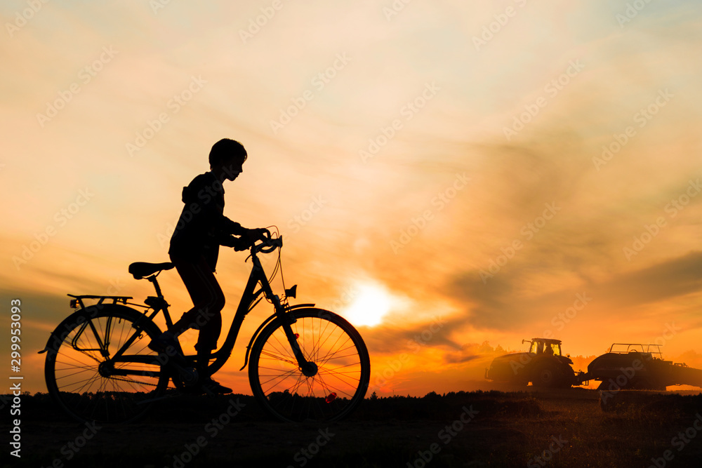 Boy , kid 10 years old riding bike in countryside, tractor working in background,  silhouette of riding person and machine at sunset in nature