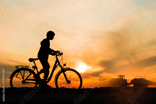 Boy , kid 10 years old riding bike in countryside, tractor working in background, silhouette of riding person and machine at sunset in nature