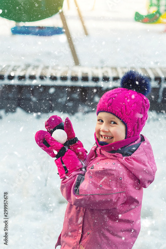 Cute adorable caucasian little girl winter portrait holding snowball in hands ready for snow fight at playground outdoor. Funny playful child during snowfall at cold season outside. Happy childhood