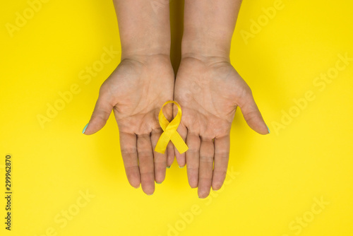 Yellow gold ribbon in the hands of a girl on a yellow background. Children cancer concept. Awareness month. Childhood Cancer Day February, 15