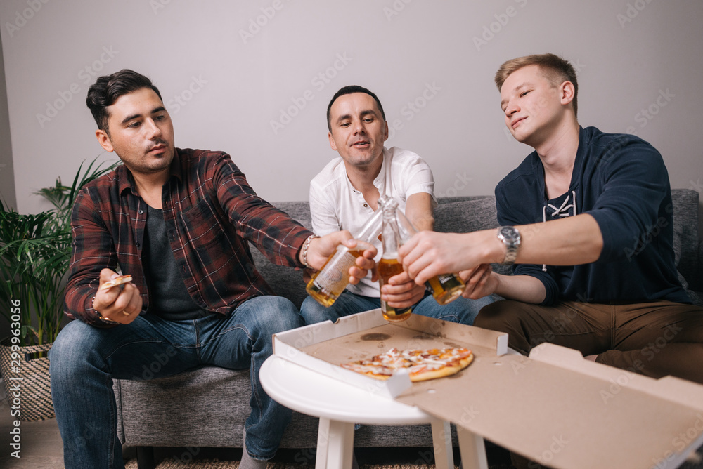 Three handsome young friends eating pizza taking it from table. Three cheerful guys are holding alcohol bottles in their hands and clinking beer bottles.