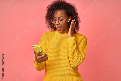 Fotografia Overjoyed happy excited Afro American female glad to answers the video call using wireless earphones, rejoices good news, stares at the mobile phone, celebrates her success