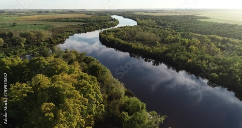 Aerial of river with natural vegetation at riverbends and reflection of sky over water surface. Agricultural landscape and sunset at background. Dolores, Soriano, Uruguay