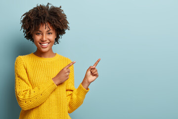 Cheerful Afro woman points away on copy space, discusses amazing promo, gives way or direction, wears yellow warm sweater, has pleasant smile, feels optimistic, isolated over blue background