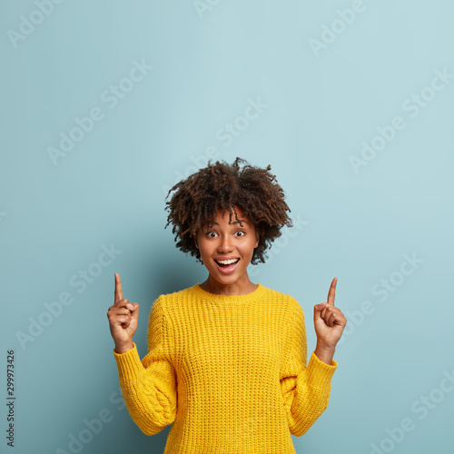 Obraz na plátně Gorgeous cheerful smiling woman with Afro hairstyle, points up, shows cool promo