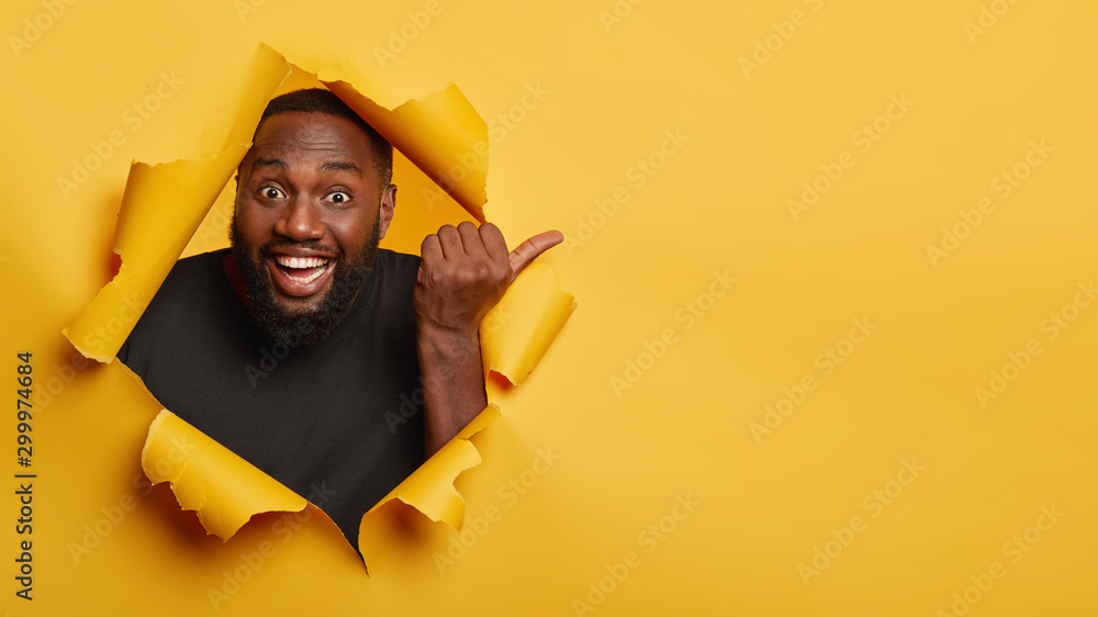 Cheerful enthusiastic Afro man with dark skin points thumb aside, laughs and introduces promo, recommends best offer, dressed in black clothing, poses through torn paper hole, yellow background - obrazy, fototapety, plakaty 