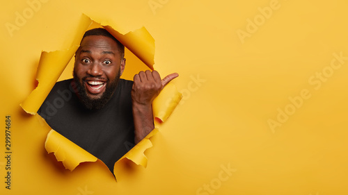Cheerful enthusiastic Afro man with dark skin points thumb aside, laughs and introduces promo, recommends best offer, dressed in black clothing, poses through torn paper hole, yellow background