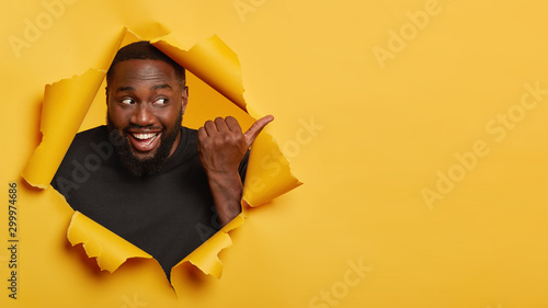 Pleased unshaven Afro American plump guy with dark skin, points thumb away, shows copy space for advertisement, pleasant promo, smiles broadly, wears black t shirt, stands in ripped paper hole © Wayhome Studio