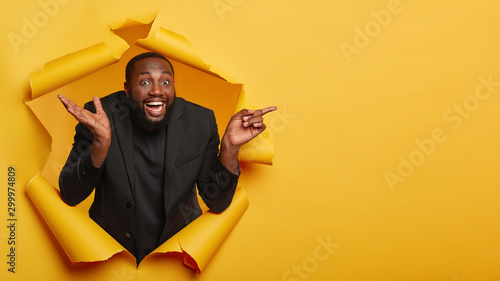 Cheerful enthusiastic man with black skin, raises palm, points away, smiles and being amused, shows amazing cool offer, dressed in formal wear, stands in ripped paper hole of yellow background © Wayhome Studio