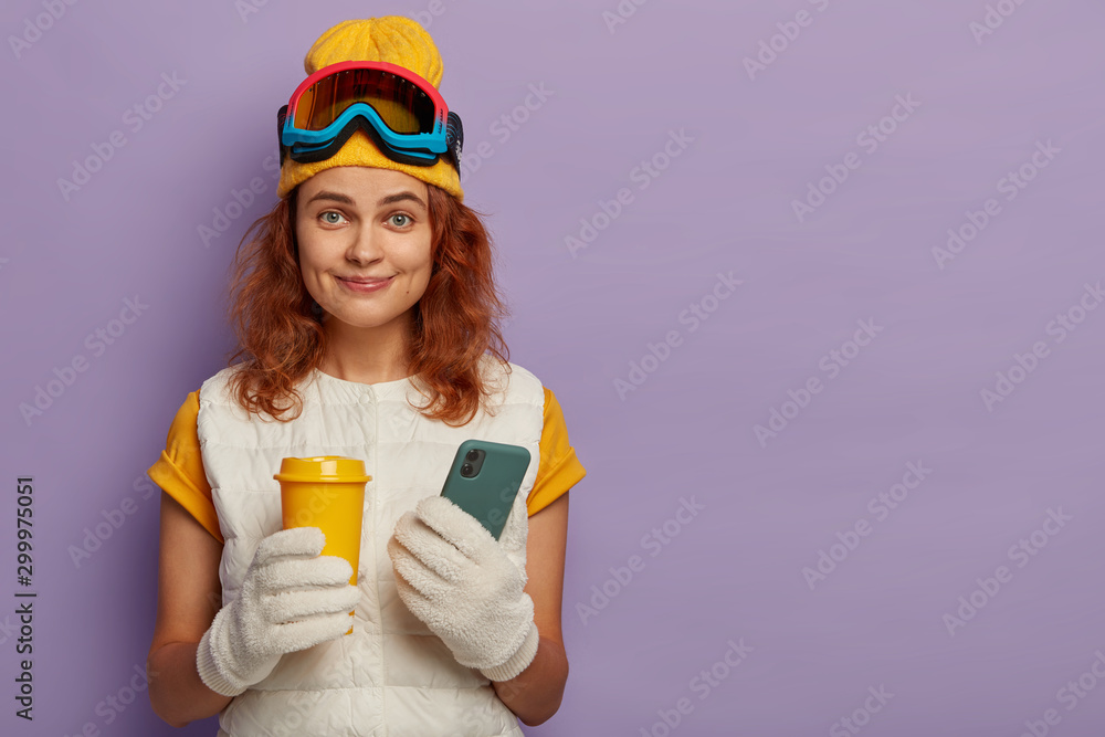 Extreme winter sports, recreation and technology concept. Happy redhead woman holds takeaway coffee and modern mobile phone, being professional skier, posts photos in social networks. Ski resort