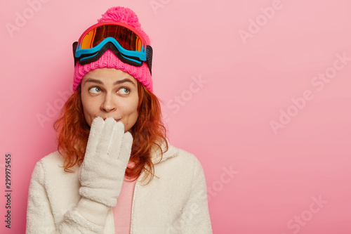 Portrait of snowboarder woman has thoughtful expression, keeps hand on mouth, wears winter hat, white gloves and ski mask, imagines nice trip to mountains, goes in for sport durimg cold weather