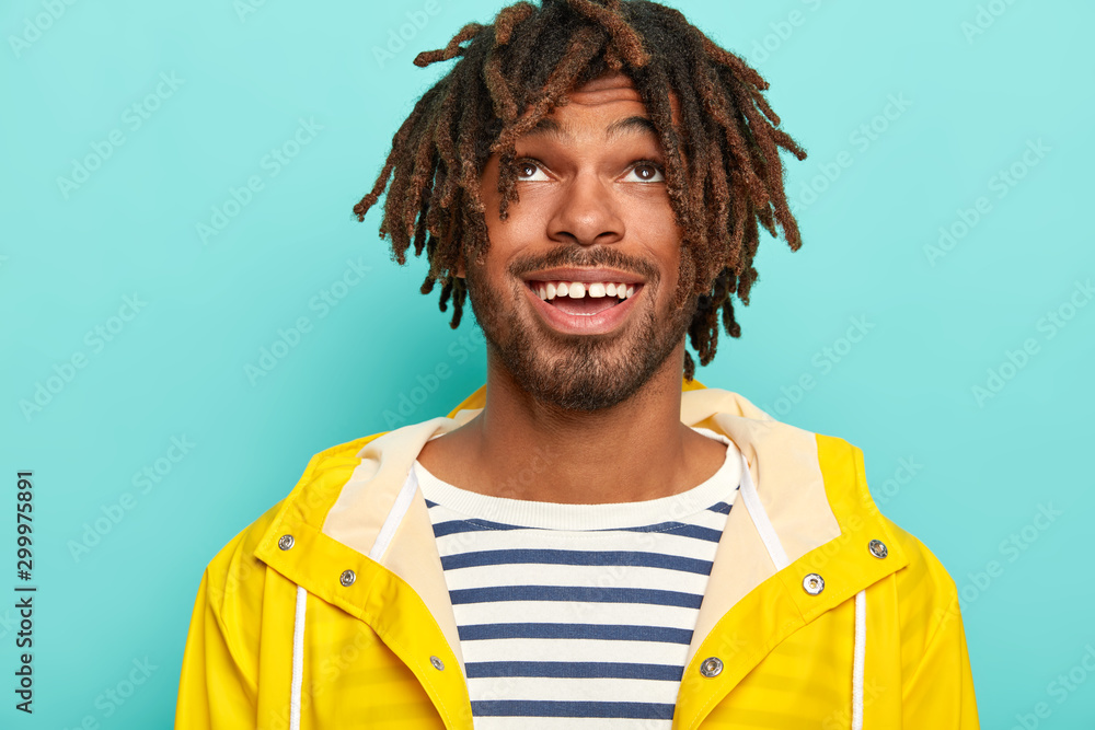 Headshot of happy man with dreads, has positive expression, focused above, wears sailor jumper and yellow raincoat, isolated over blue background, enjoys rainy autumn weather. Cheerful teenager Headshot of happy man with dreads, has positive expression, focused above, wears sailor jumper and yellow raincoat, isolated over blue background, enjoys rainy autumn weather. Cheerful teenager