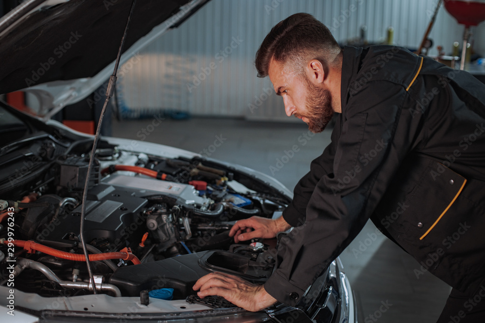 A young mechanic is very focused on looking at the engine of the car he has to repair A young mechanic is very focused on looking at the engine of the car he has to repair
