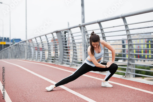 City fitness. A young sports woman in fitness suit is warming up, doing stretching against steel railing of the bridge.