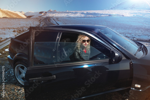 Fashion style wide angle young stylish attractive girl with bright red lips in a leather jacket and sunglasses sits in a retro 90s car with an open door on nature in the mountains at sunset