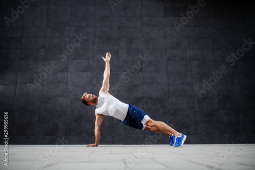 Handsome sporty caucasian man in shorts and t-shirt stretching his arm in plank position in front of gray wall.