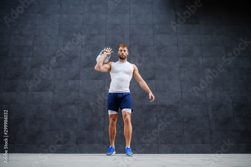 Full length of handsome strong muscular bearded Caucasian man in shorts and t-shirt lifting kettle bell while standing outdoors in front of gray wall.