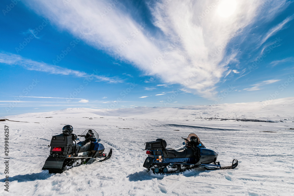 Two blue snowmobile stands on snow in Norwegian mountains, warm day and bright sun, snow smelts, norwegian mountains landscape, back side view, black safety helmets lay at snowmobiles