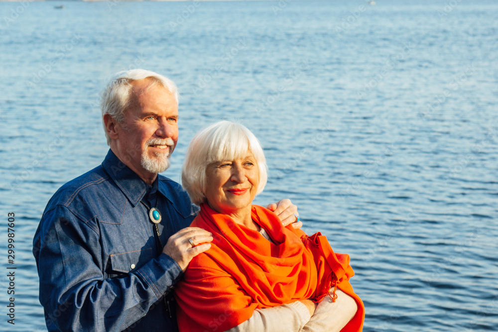 cheerful senior citizens woman and man are standing and hugging on the lake, against the background of the bridge.
