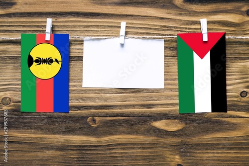 Hanging flags of New Caledonia and Palestine attached to rope with clothes pins with copy space on white note paper on wooden background.Diplomatic relations between countries.