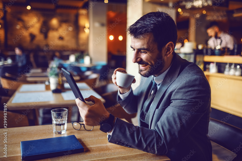 Handsome smiling caucasian bearded businessman in suit sitting in cafe, drinking coffee and using tablet. - obrazy, fototapety, plakaty 