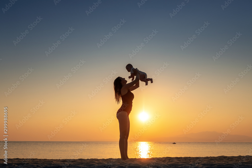 Silhouette of committed mother in swimsuit lifting her loving 6 months old baby boy while standing on the beach.