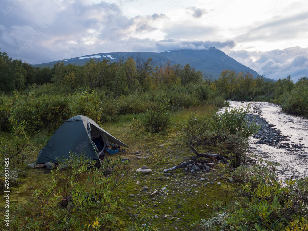 Small green tent in Beautiful wild Lapland nature landscape with blue river, birch trees and mountains. Northern Sweden summer at Kungsleden hiking trail. Blue sky dramatic clouds